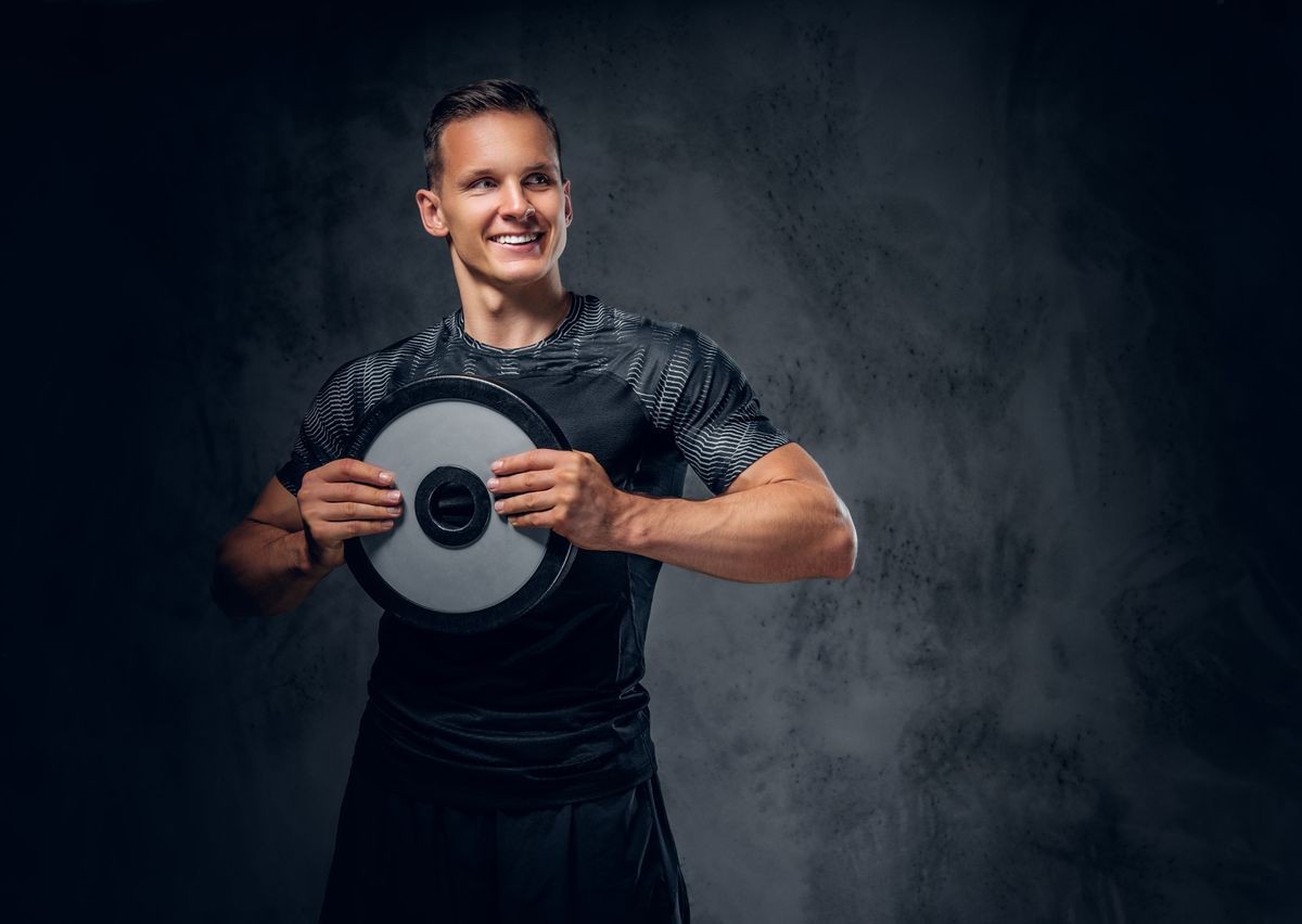 Athletic male holds a barbell weight in his arm over grey background.