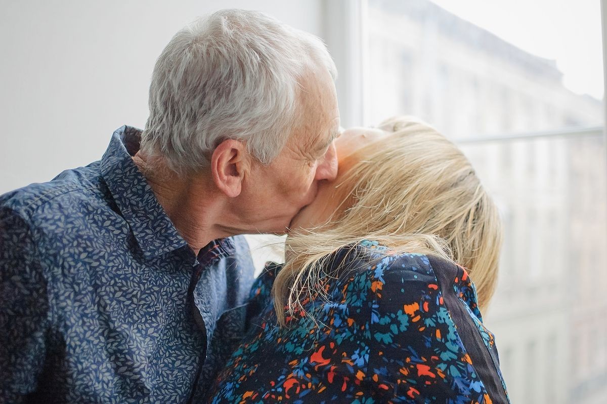 Hot and Sexy Middle-aged Woman Enjoying Kissing of Her Elderly Husband Standing near Opened Window inside Their Home. Couple with Age Difference